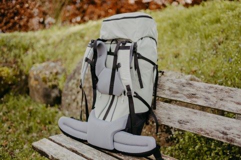 Un grand sac à dos de trek gris posé sur un banc en bois dans la verdure.