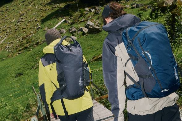 Zwei Wanderer von hinten mit Rucksäcken auf dem Rücken laufen über eine Holzbrücke