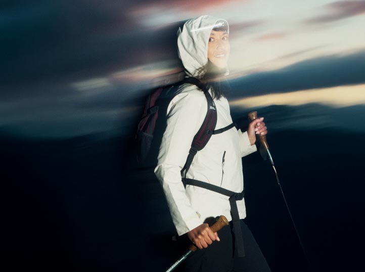 Une personne en vêtements de randonnée, incluant une veste blanche avec capuche, tenant des bâtons de trek. Le ciel flou du crépuscule suggère le mouvement et l'aventure.