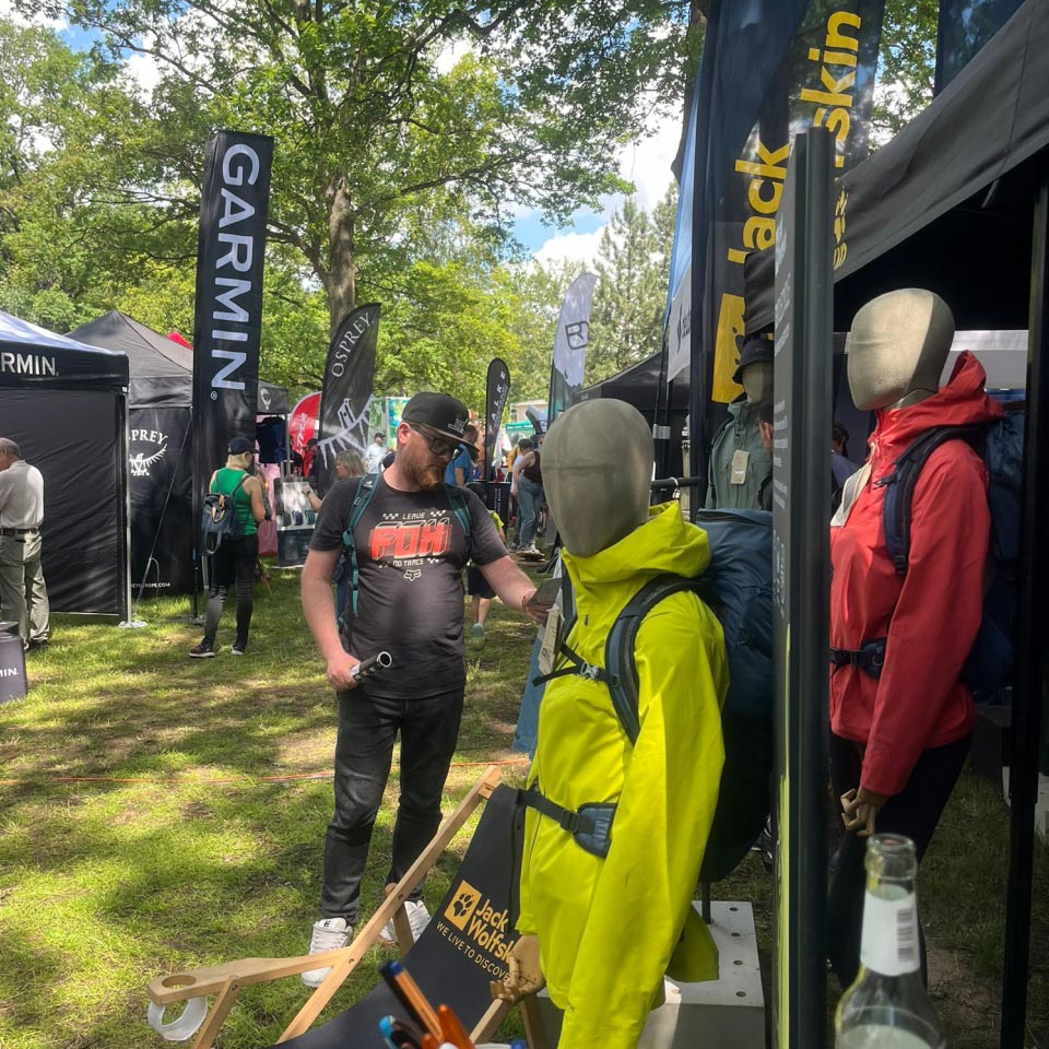 Mann in schwarzem Shirt steht vor einem Stand mit Outdoor-Bekleidung und Markenflaggen im Hintergrund