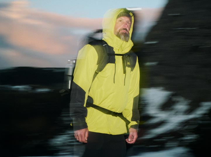 Un homme avec une veste jaune vif et un sac à dos en train de randonner au crépuscule ; le mouvement flouté confère à l'image un caractère dynamique.