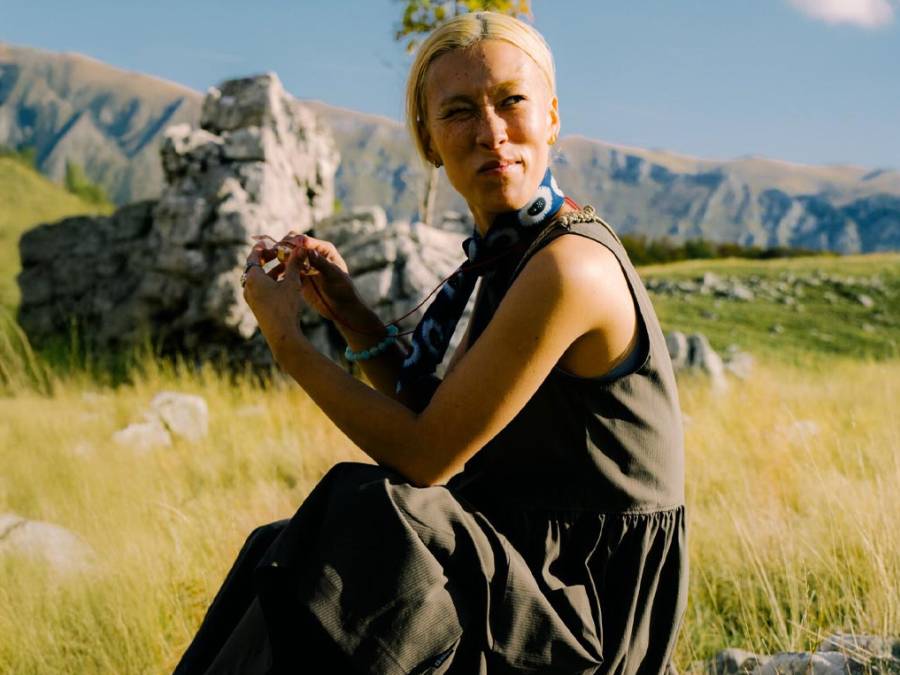 Eine blonde Frau sitzt in einer grünen Wiese und schaut in die Ferne, umgeben von Bergen