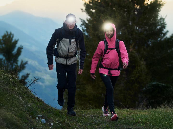 Deux randonneurs avec des lampes frontales marchent au crépuscule sur un chemin forestier. Ils portent des vestes, l’une noire et grise, l’autre rose.