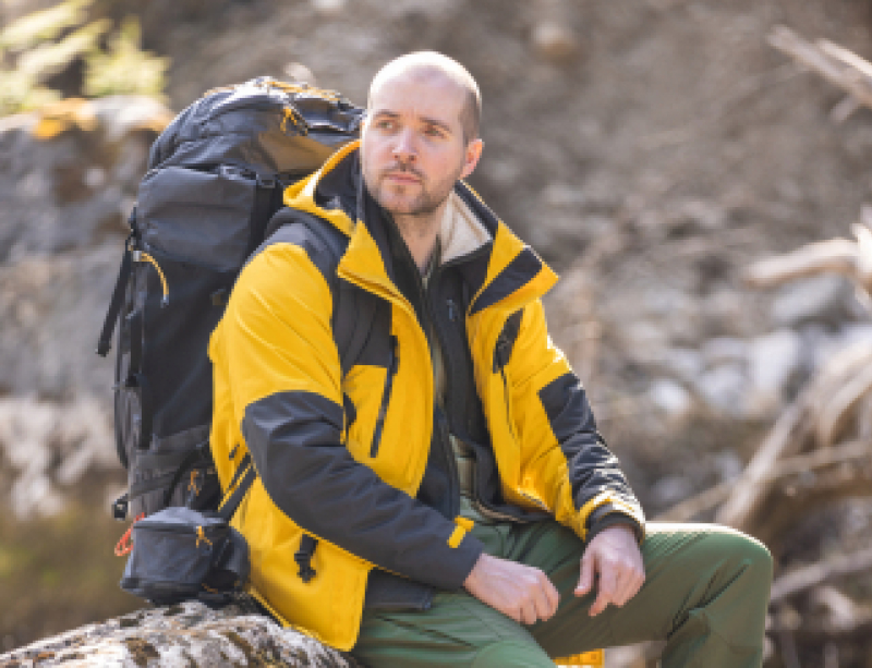 Chris sitzt mit Wanderrucksack auf einem Baumstamm